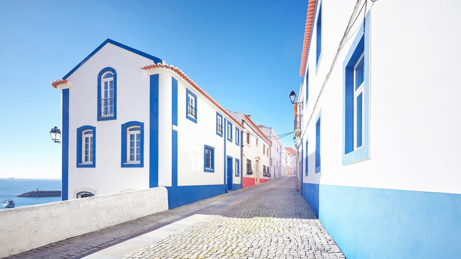 Solrik gate med tradisjonell portugisisk arkitektur, brosteinbelagt gate og et glimt av havet.