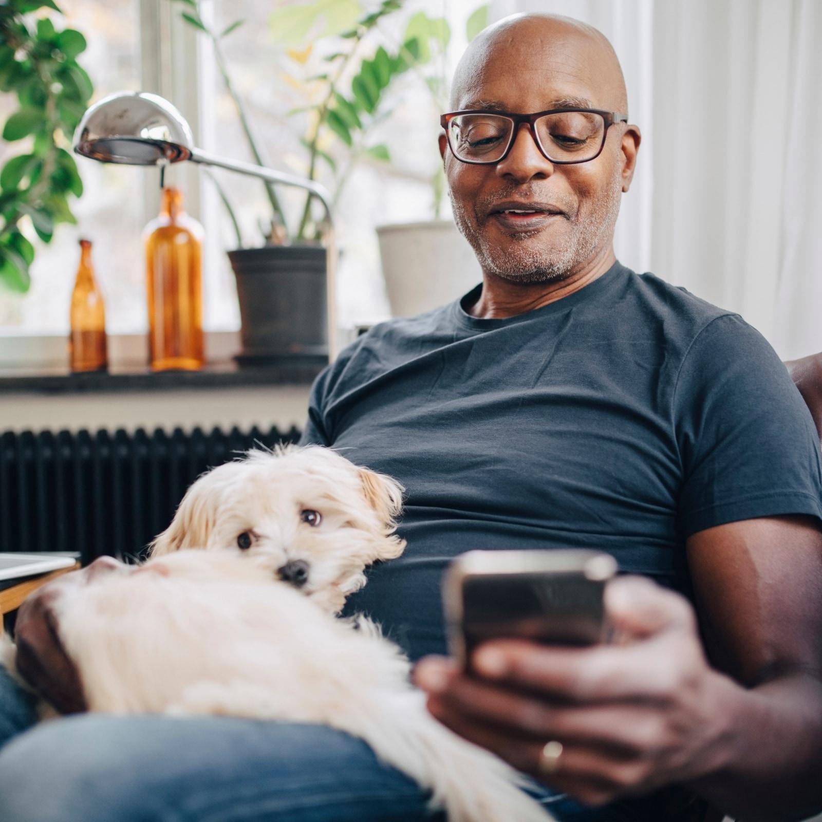 Homem de óculos e camisa azul a relaxar no sofá com um cão pequeno e peludo, a olhar para o seu smartphone.